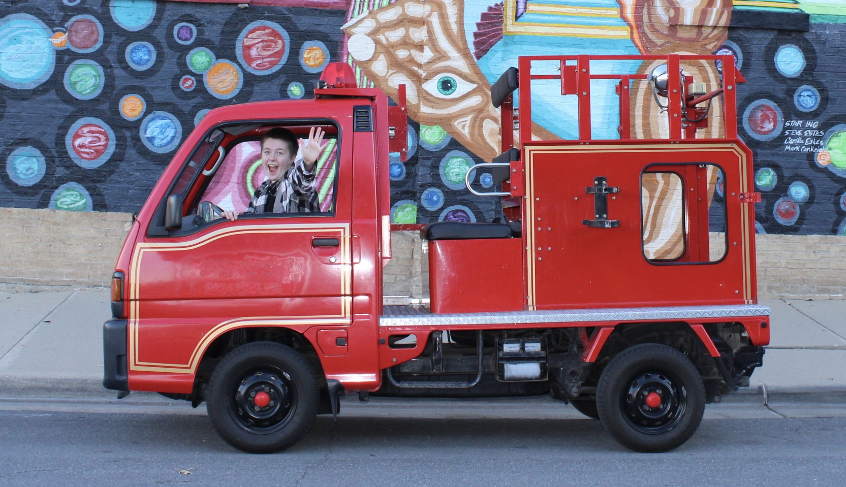 Jordan waving from the driver's seat of a small red fire truck parked in front of a colorful mural.