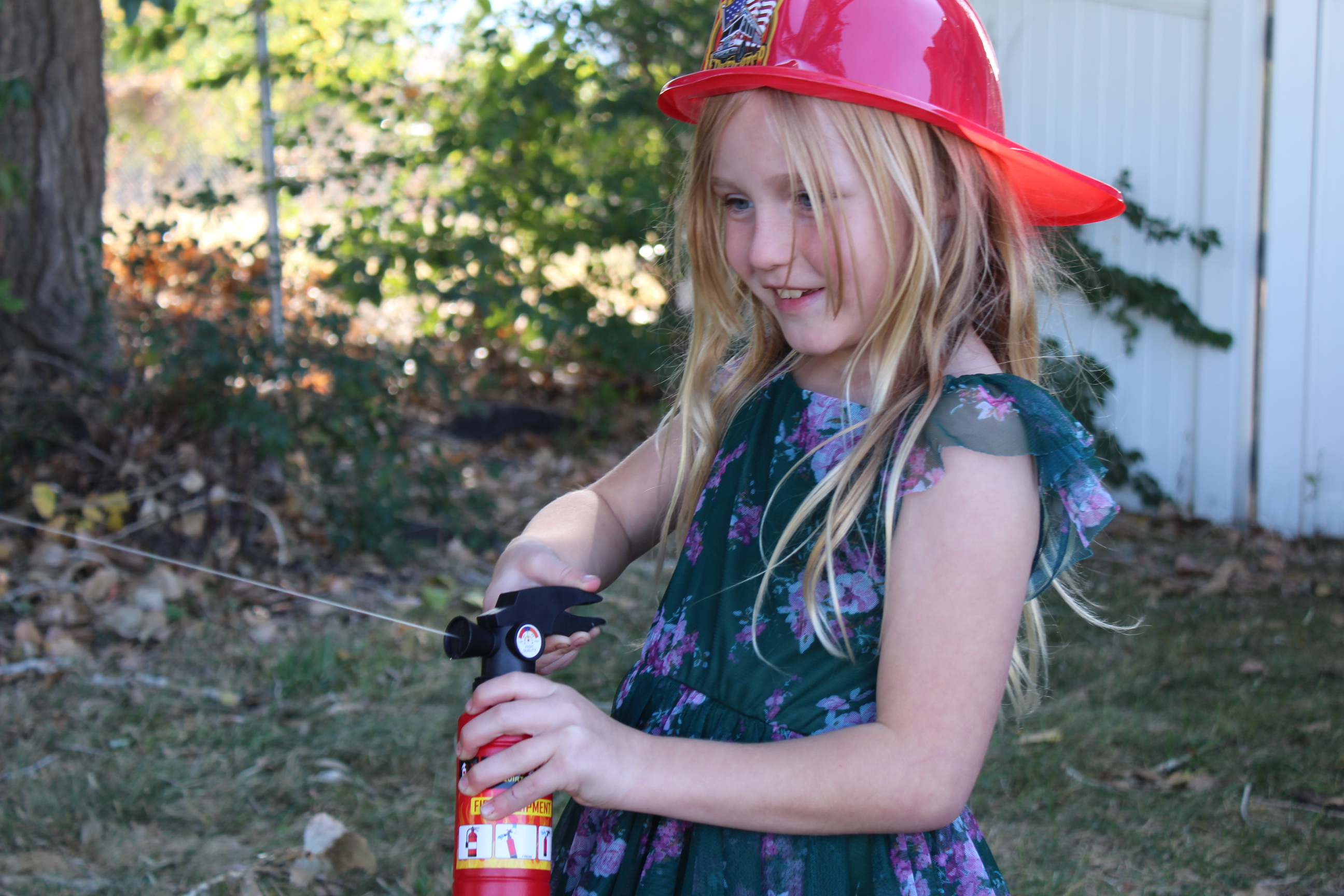 A young girl with long blonde hair smiles while playing outdoors. She wears a bright red firefighter helmet and a green floral dress. In her hands, she holds what appears to be a toy fire extinguisher, pretending to be a firefighter.