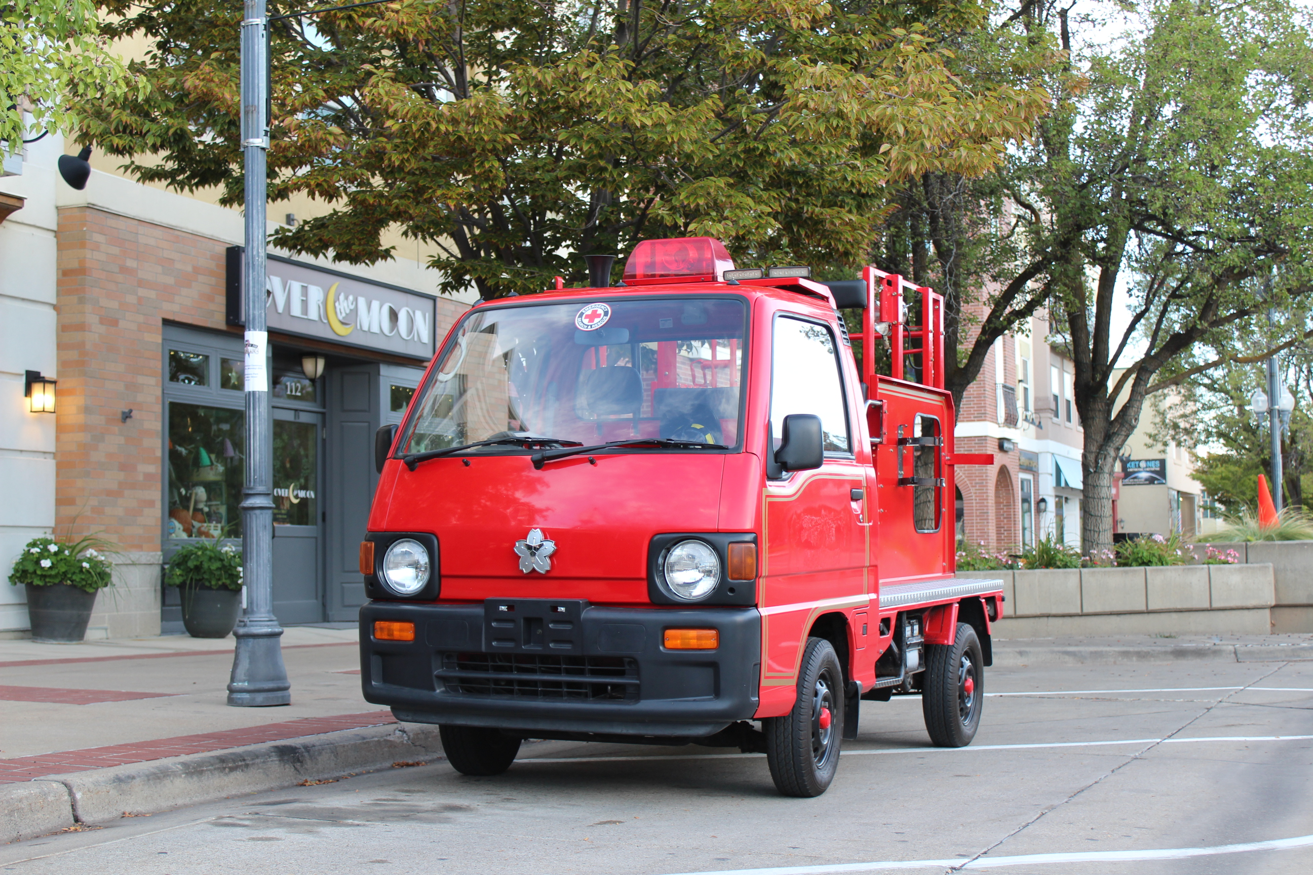 A small red fire truck parked on a city street. The compact vehicle has a flat front with round headlights and a utility bed in the back. It's parked in front of a shop called 'MH & MOON' with trees lining the street. The scene appears to be in a quaint downtown area with brick buildings and planters on the sidewalk.