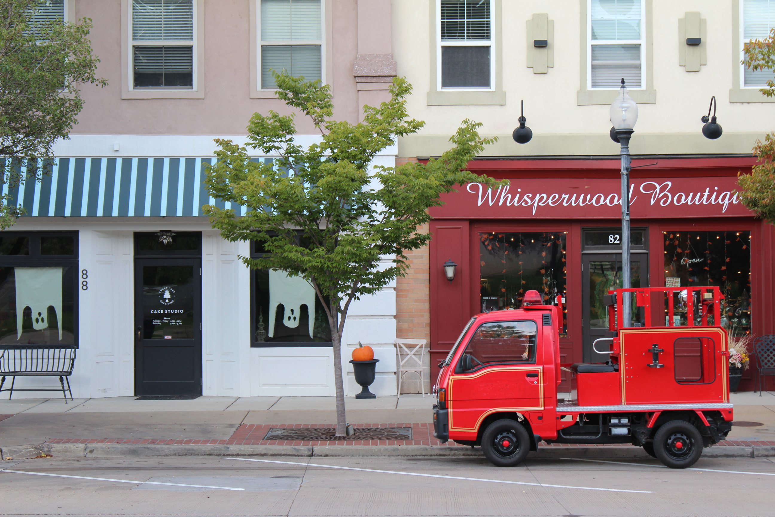 A small red fire truck parked in front of two shops on a city sidewalk. On the left is a store with a blue and white striped awning and ghost decorations in the window. On the right is 'Whisperwood Boutique' with a red storefront. Trees line the street.