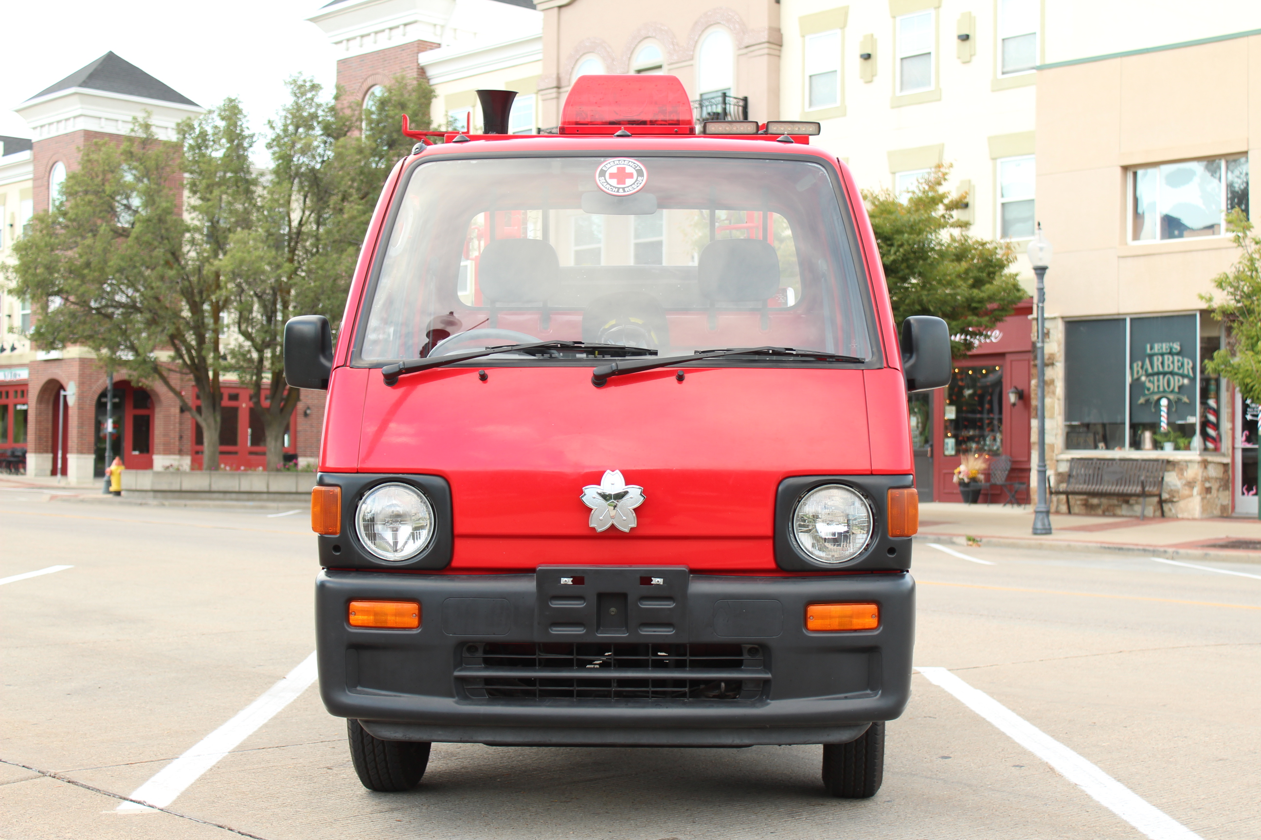 Front view of a small red fire truck parked on a city street. The vehicle has round headlights and a silver emblem on its grille. Behind it are colorful buildings including storefronts and what appears to be a barber shop.