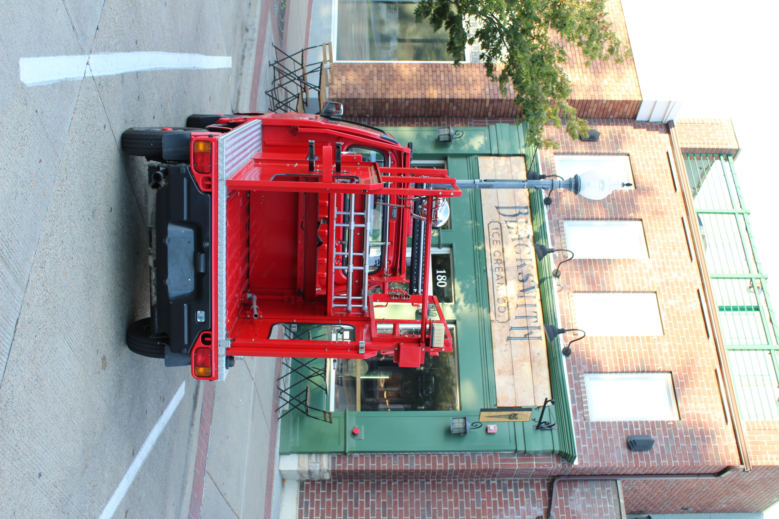 Rear view of a small red fire truck parked in front of 'Blacksmith Ice Cream Co.'. The truck's utility bed is visible, featuring red metal frameworks. The ice cream shop has a green storefront with outdoor seating.