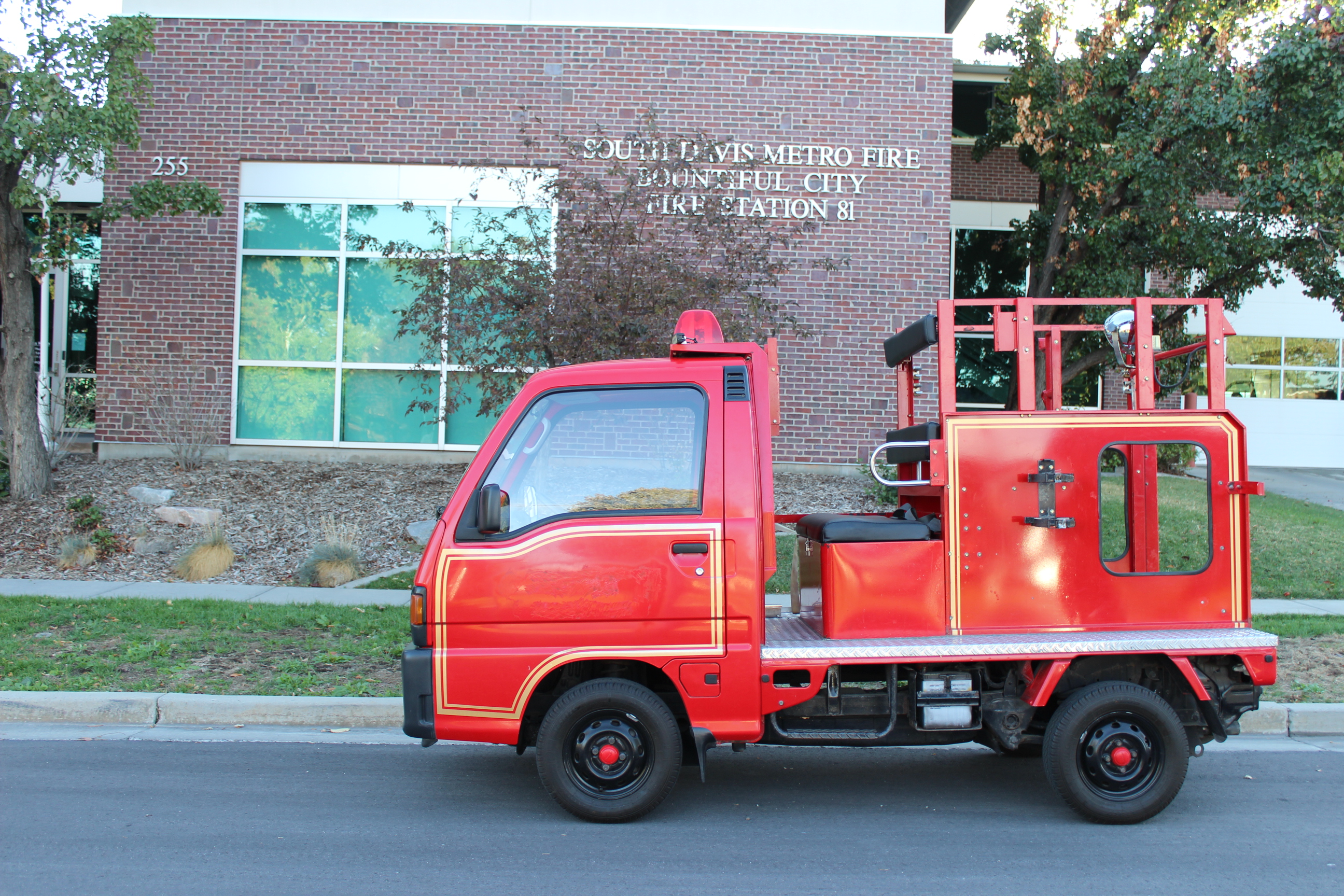 A small red fire truck parked in front of the South Davis Metro Fire Station 81 in Bountiful City. The fire station is a brick building with large windows. The compact fire truck has equipment mounted on its flatbed.