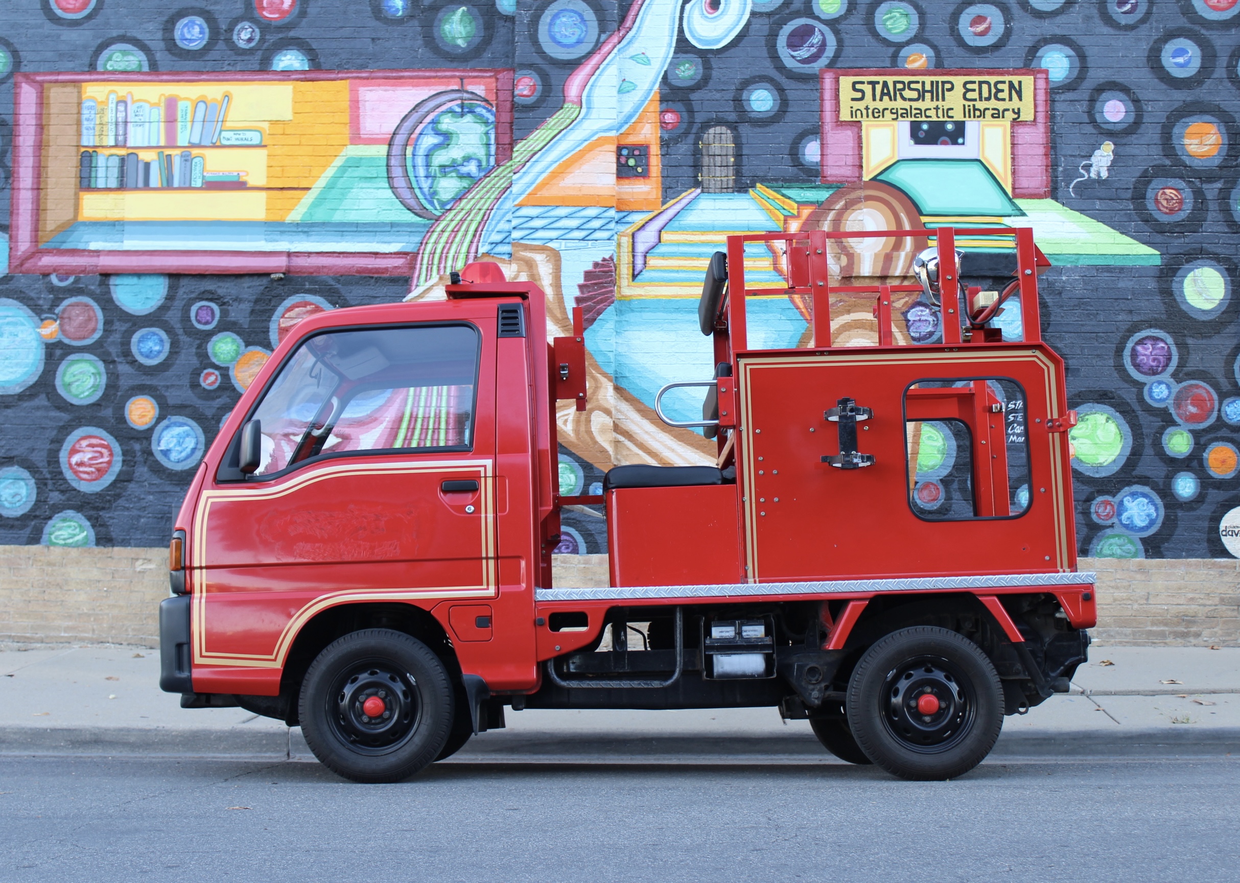 A small red fire truck parked in front of a colorful mural. The mural depicts a fantastical library scene with the text 'STARSHIP EDEN intergalactic library' visible. The fire truck has a compact cab and a utility bed with equipment. The background is filled with vibrant, cartoon-like circular designs resembling planets or bubbles.