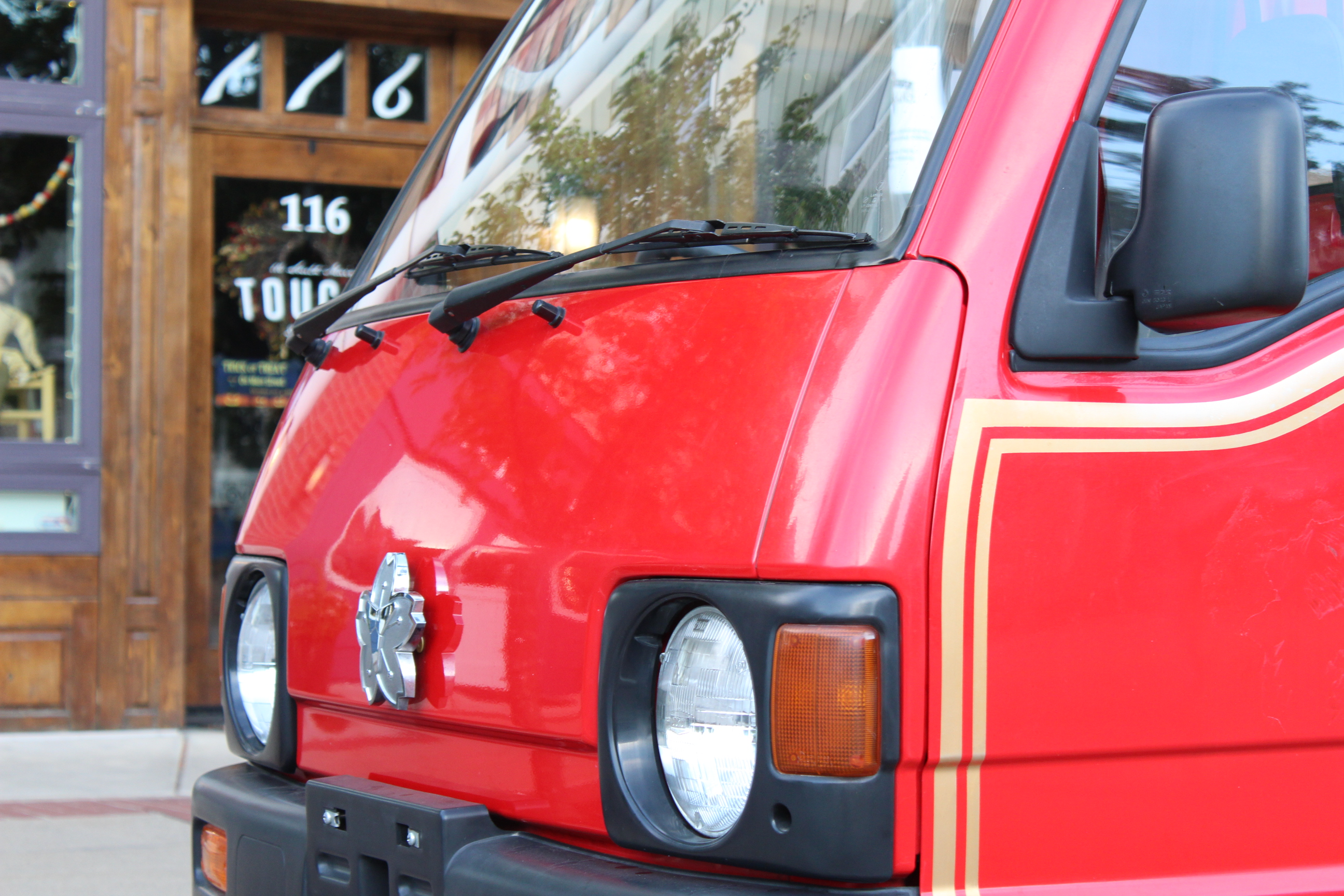 Close-up view of the front of a small red fire truck. The image shows the truck's round headlights, turn signal, and a silver emblem on the grille. A yellow pinstripe decorates the side. In the background, a wooden storefront with the number 116 is visible, along with part of a sign reading 'TOUS'.